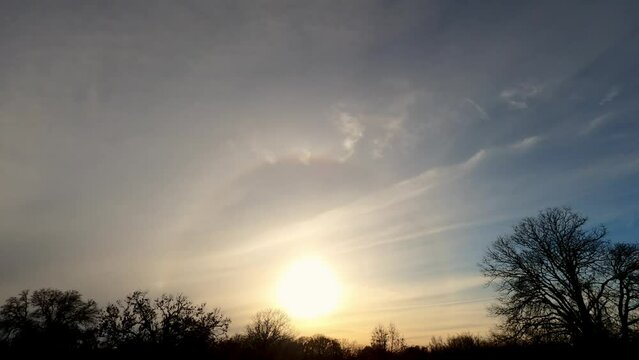 Timelapse of a halo with sun dogs around setting sun in thin clouds in winter; over rural landscape