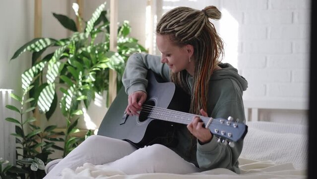 woman with dreadlocks on head singing song and playing acoustic guitar in sunny cottage bedroom, simple living and hobby relaxing