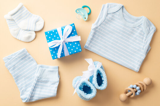 Baby boy concept. Top view photo of blue giftbox with bow infant clothes shirt panties knitted booties socks pacifier and wooden rattle on isolated pastel beige background