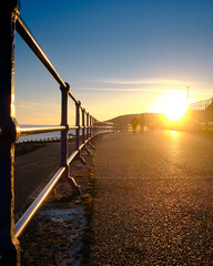 Blue railings on Eastbourne promenade with clear sunset sky. Pebbled beach with wooden groynes and a calm sea.