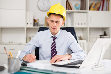 Male engineer sitting in an office, working at a computer and making important notes