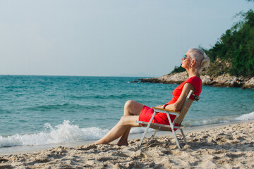 Happy Caucasian senior woman sitting on beach chair on the beach over sunset. Retirement elderly female resting by the sea relax and enjoy outdoor lifestyle activity in summer vacation