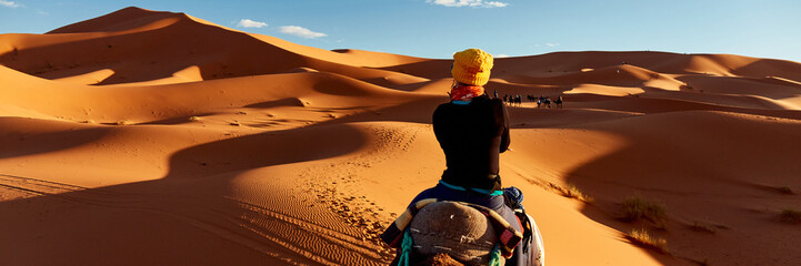 A young women in a yellow cap rides a camel  through the dunes in the Sahara Desert. View of the woman from behind, in the background, small silhouettes of other tourists. Merzouga, Morocco - obrazy, fototapety, plakaty