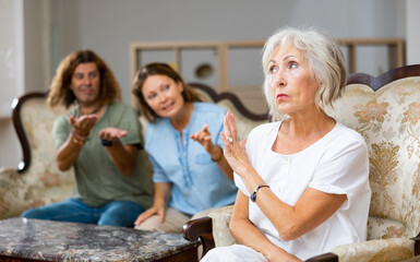 Dissatisfied mother-in-law siting on a chair against of a confused husband and wife on the sofa in...