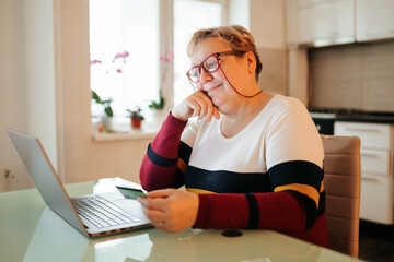 A mature, overweight woman, who is feeling pleased and positive, is shown shopping online and paying with her credit card from the comfort of her own home. - obrazy, fototapety, plakaty