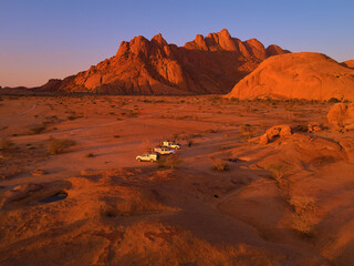 Fototapeta na wymiar Aerial view of campsite at the foot of Spitzkoppe. Three offroad camper cars on campsite against red, bald granite peaks illuminated by setting sun. Travelling Namib desert, Namibia. 