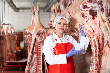 Positive young female butcher working in chilling room of meat processing factory, arranging raw...