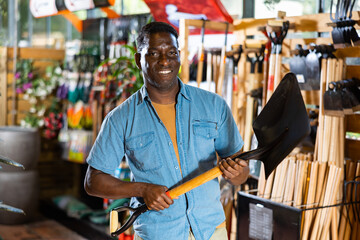 Portrait of smiling adult african american gardener buying shovel in gardening market..