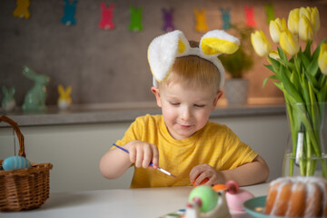 A little boy wearing bunny ears prepares painted Easter eggs for Easter while sitting at the kitchen table. Happy easter.