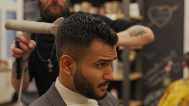 Professional male barber arranges his indian client's hair. He uses a brush and a dryer for this. High quality 4k footage