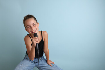 Portrait of a cute teenage girl singing, using microphone. Teenage girl in black t-shirt on an isolated blue background. Karaoke for kids, home entertainment for kids. Singer singing with microphone.
