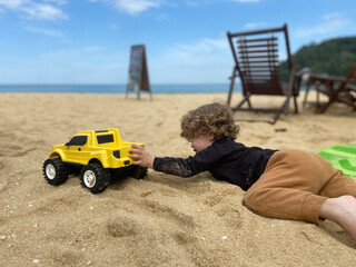 blonde child dressed in pants and a long-sleeved shirt lying on the fine sand playing with her toy yellow pickup truck on a sunny summer day