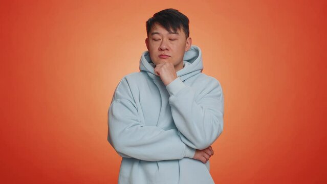 Thoughtful clever asian handsome man rubbing his chin and looking aside with pensive expression, pondering a solution, doubting question. Adult guy on orange studio background. People lifestyles