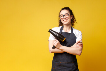young girl hairdresser in uniform holds a hair dryer and smiles on a yellow background, woman...