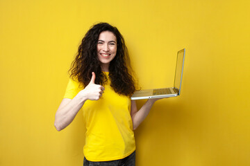 young curly woman in a yellow t-shirt holds a laptop and shows like with her hand on yellow...