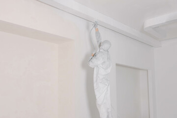 Decorator in protective overalls painting wall with spray gun indoors