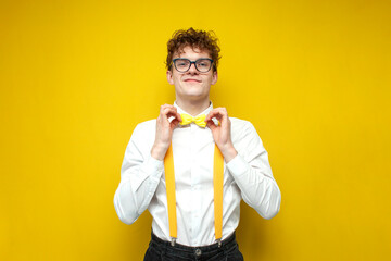 young curly guy in shirt and suspenders straightens his bow tie on yellow background, smart nerd in...