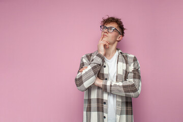 pensive curly guy in glasses stands on a pink background and thinks, a man in a plaid shirt dreams and looks away