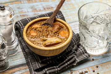 Traditional Spanish lentil stew inside a wooden bamboo bowl on a black and gray kitchen towel, fresh tomatoes, a wooden spoon and cut crystal glasses on a blue wooden table