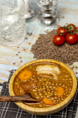 Traditional Spanish lentil stew inside a wooden bamboo bowl on a black and gray kitchen towel, fresh tomatoes, a wooden spoon and cut crystal glasses