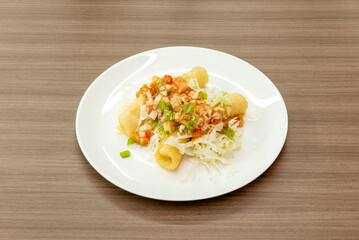 A bright white plate of Mexican flautas with minced meat stew and salad on a light brown table