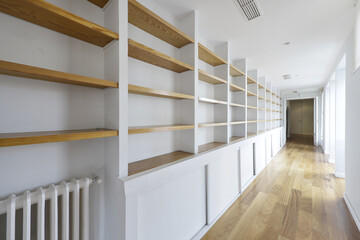 Long hallway of a home with a long empty bookcase with wooden shelving and a loose oak floor