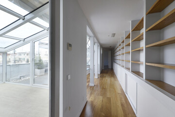 Long corridor of a penthouse apartment with a glazed terrace and a long empty shelf with wooden shelves and loose oak flooring