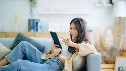 Happy young asian woman relax on comfortable couch at home texting messaging on tablet, smiling...