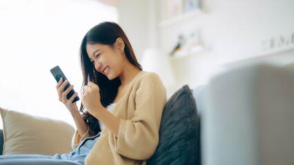 Happy young asian woman relax on comfortable couch at home texting messaging on smartphone, smiling...