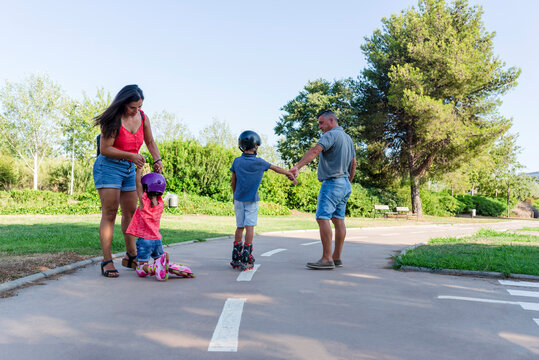 Kids holding her parents hands while roller skating outdoors in the park.