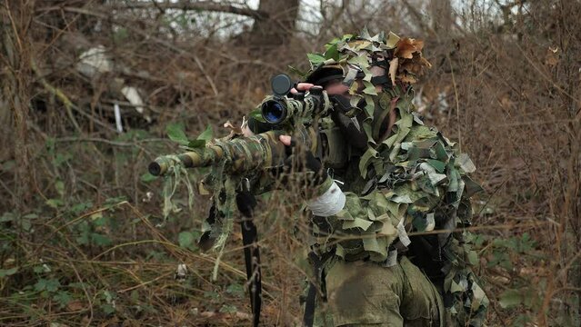 A professional military man in camouflage uniform with a sniper rifle in the forest among dry branches and grass. Portrait of a sniper in nature. War. Military conflict.
