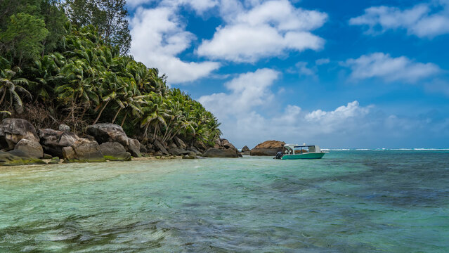 The tropical island is overgrown with lush palm trees. Boulders are piled at the water's edge. The boat is moored in the turquoise ocean. Clouds in the blue sky. Seychelles. Moyenne Island