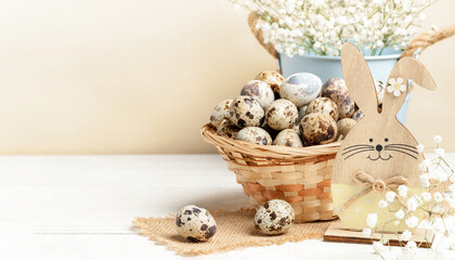 Woven basket of quail eggs, wooden bunny, white flowers on white wooden table on beige Easter card