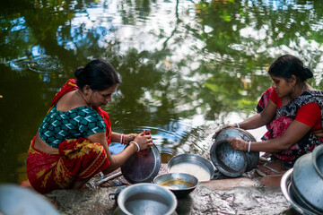 South asian rural women washing cookwares and clothes sitting beside a village pond 