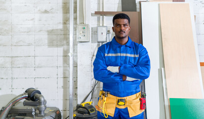 Fototapeta na wymiar Young technician in blue mechanic jumpsuit, protective glove and yellow Tool Belts stand with arms crossed in a wooden furniture factory.