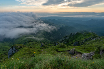 Green meadows with rocks and rocky mountains with fog in the valley and clouds in the sky in...