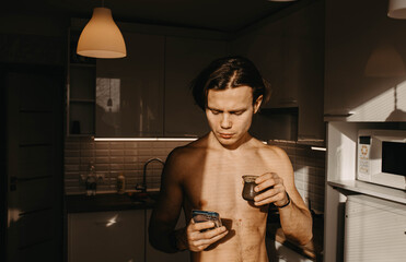 man with a naked torso drinks tea in the morning in the kitchen with a smartphone in his hands.