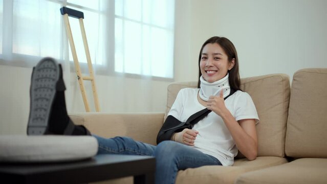 Happy woman recovery from accident fracture broken bone injury with leg splints in cast, neck splints collar, arm splints, sling support arm in living room. Social security and health insurance.