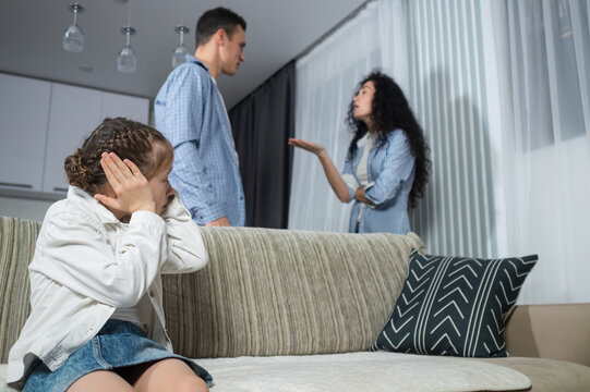 Junior schoolgirl sitsclosing ears with hands and looking at parents against arguing behind sofa. Angry father and mother quarrel and argue about daughter naughty behavior