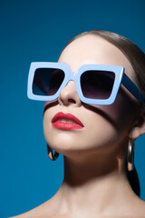 Fashion and make-up concept. Close-up beautiful woman portrait with classic hairstyle, big retro blue sunglasses, red lipstick and golden round earrings in blue studio background. Model looking up