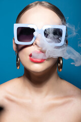 Naklejka premium Fashion and make-up concept. Woman portrait with classic hairstyle, big retro sunglasses, red lipstick and golden round earrings blowing white dense smoke from mouth in blue studio background