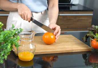 Closeup hand holding knife cutting orange fruit on a wooden chop board. Jar with mixed fruit juice  placed on the kitchen counter.