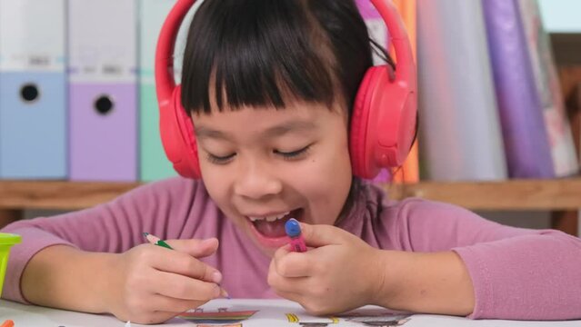 Cheerful little girl in headphones singing and drawing with colored pencils on paper sitting at table in her roCheerful little girl in headphones singing and drawing with colored pencils on paper sitt