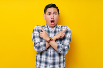 Shocked Young Asian man in casual clothes crossed arms and points sideways, chooses between two options, indicates left and right isolated on yellow background. People lifestyle concept