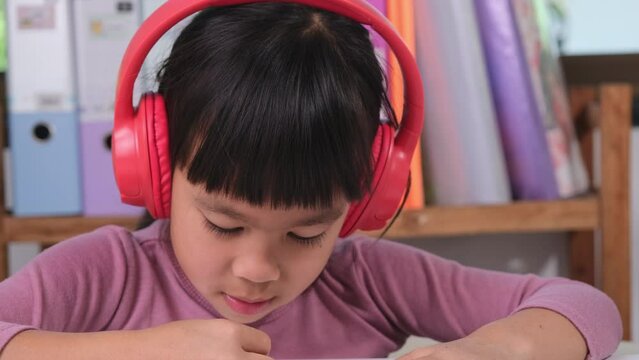 Cheerful little girl in headphones singing and drawing with colored pencils on paper sitting at table in her room at home. Creativity and development of fine motor skills.