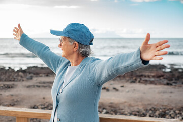 Smiling senior woman in sea vacation standing outdoors at beach in sunset light with outstretched arms looking at horizon. Happy pensioner woman expressing freedom and positivity