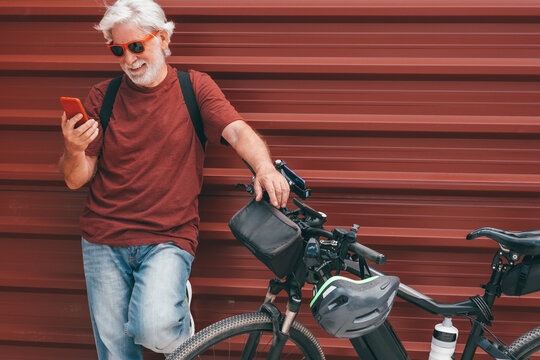 Handsome senior man standing near his electric bicycle in the city wearing sunglasses using mobile phone against red metal background. Old man with white hair and beard appreciates modern technology