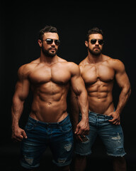 Fototapeta na wymiar Two shirtless hunks at black background. Fitness models couple in jeans and sunglasses. Two muscular handsome men with six pack abs in studio.