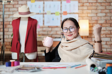 Asian mature female fashion designer who smiles and looks at camera celebrates and is cheerful with dress design work success and joyful in stylist studio, a happy costume boutique small business.