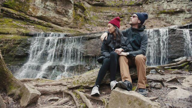 Man and woman sit on rocks at waterfall 
